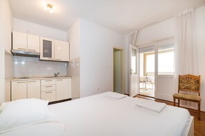 A bedroom with a bed, a wooden chair, a kitchenette with white cabinets, and a door leading to a balcony.