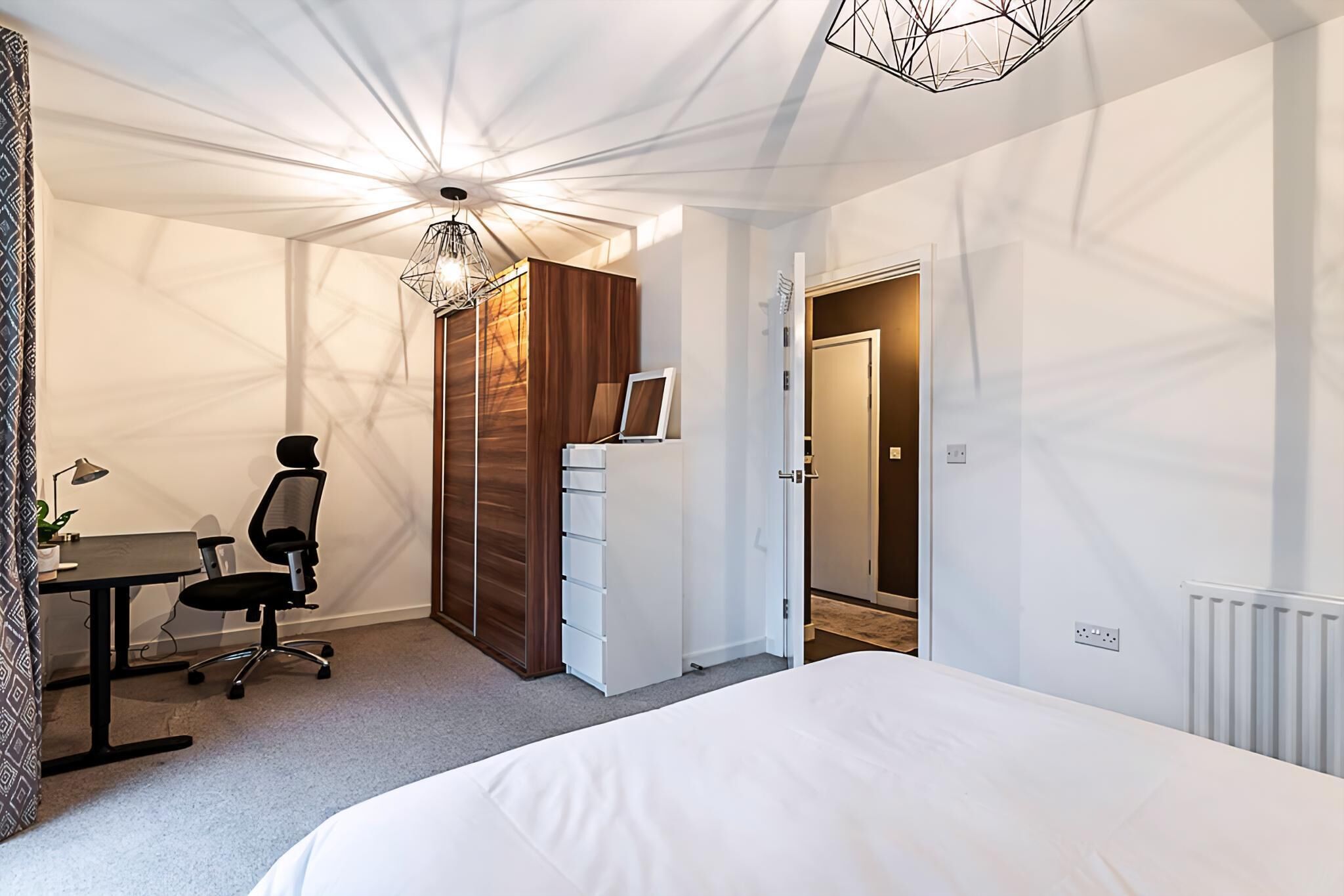 A modern bedroom with a large bed, a desk with a chair, a wardrobe, and a ceiling light fixture.