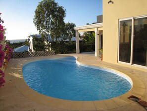 A swimming pool with a curved edge, a covered patio area, and a view of a building with large windows.
