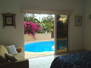 Quarto com cama, sofá, porta de vidro que se abre para uma piscina e vista para o verde lá fora.