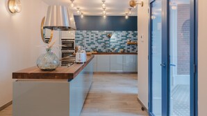 A modern kitchen with a central island, a hanging light fixture, and a view of the outside through a glass door. - Villa D'Eaux (Villers-sur-Mer)
