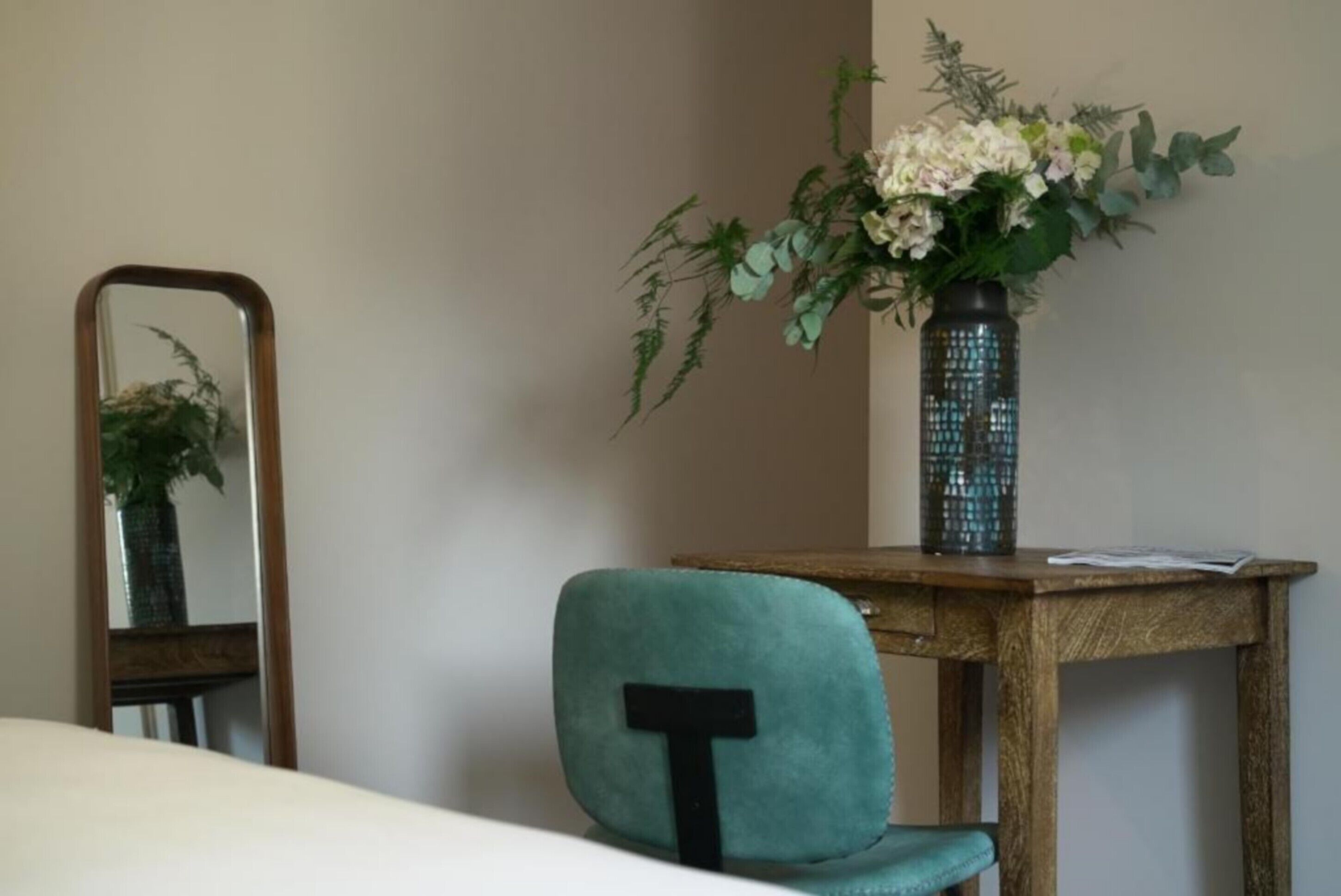 Een kamer met een smaragdgroene fauteuil, een houten tafel, een spiegel en een vaas met bloemen.