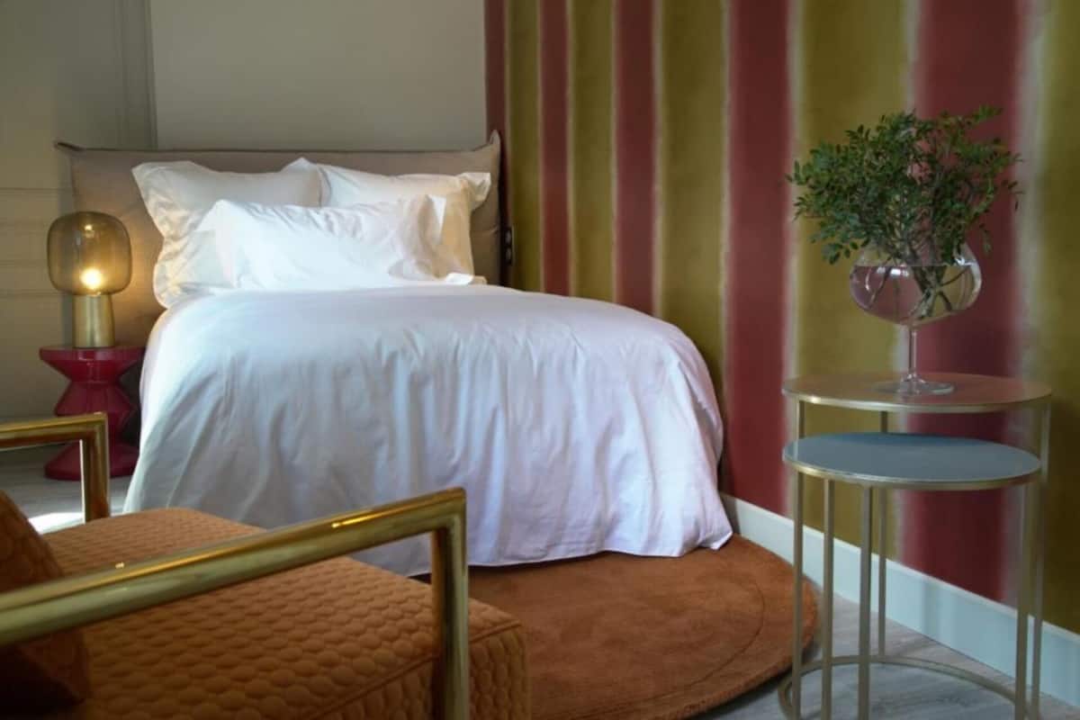 A neatly arranged bedroom with a bed, a bedside table with a lamp, a small round table with a vase of flowers, and a chair.