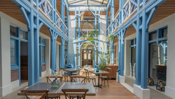 A covered outdoor area with wooden tables and chairs, blue structural elements, and potted plants. - Villa D'Eaux (Villers-sur-Mer)
