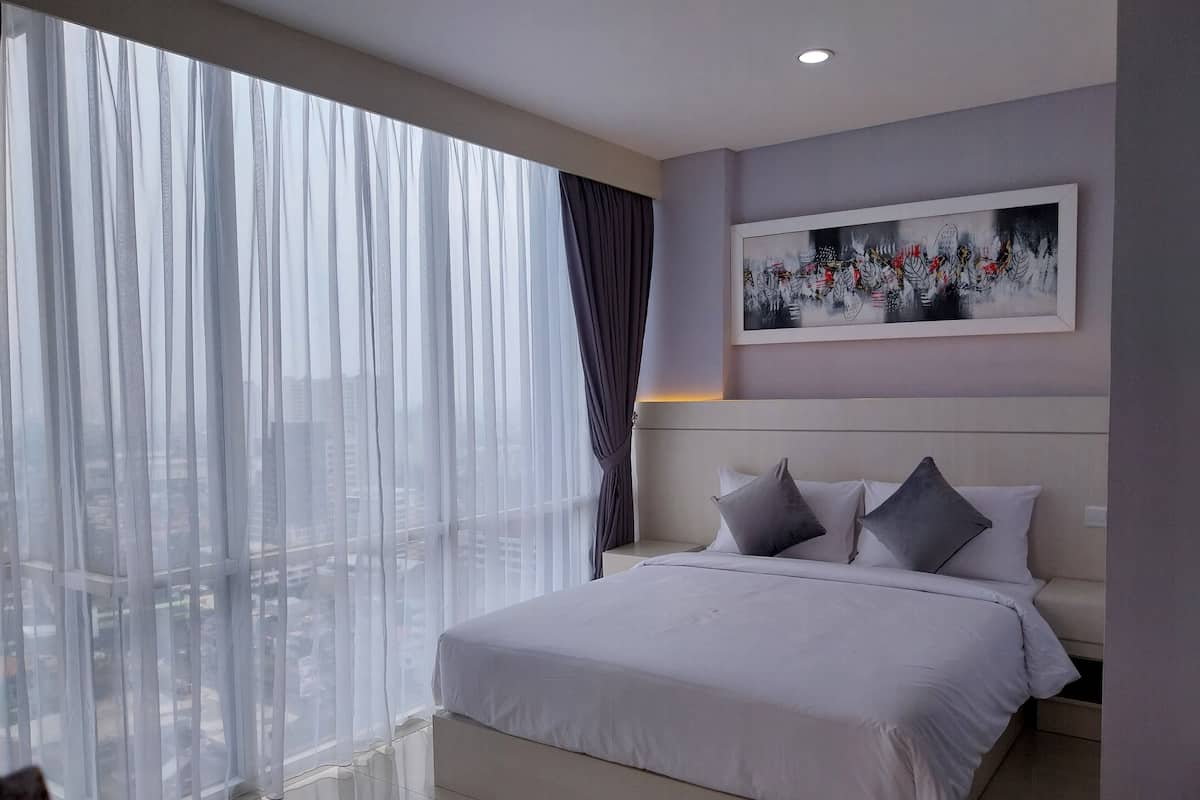 A modern hotel room with a large bed, a city view, and a framed artwork on the wall.