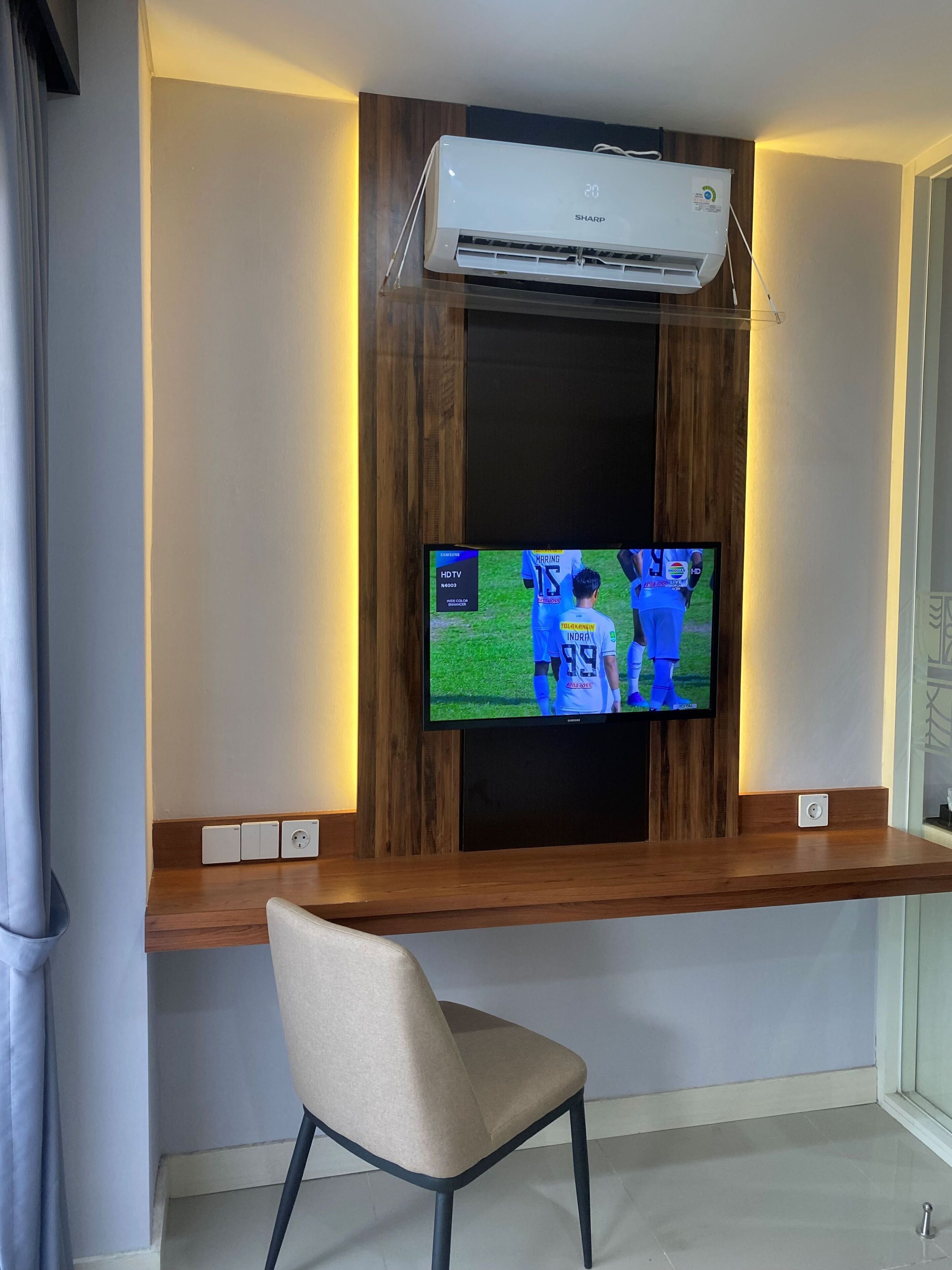 Una habitación con una televisión de montaje mostrando un evento deportivo, un escritorio de madera, una silla y un equipo de aire acondicionado sobre la televisión.