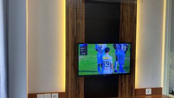 Una habitación con una televisión de montaje mostrando un evento deportivo, un escritorio de madera, una silla y un equipo de aire acondicionado sobre la televisión.