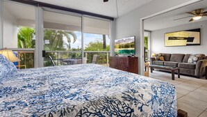 A bedroom with a large bed, a ceiling fan, a sliding glass door to a balcony, a sofa, and a coffee table.