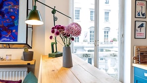 A room with a wooden table, a vase with flowers, a green lamp, a radiator, a cactus, and a city view through a window.