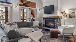Een moderne woonkamer met een open haard, een ruime bank, een aan de muur bevestigde televisie en uitzicht op buiten.