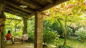 A covered outdoor seating area with stone pillars, surrounded by lush greenery and flowering plants. - Welcome Collection - Villa Mellicata (Massa Lubrense)