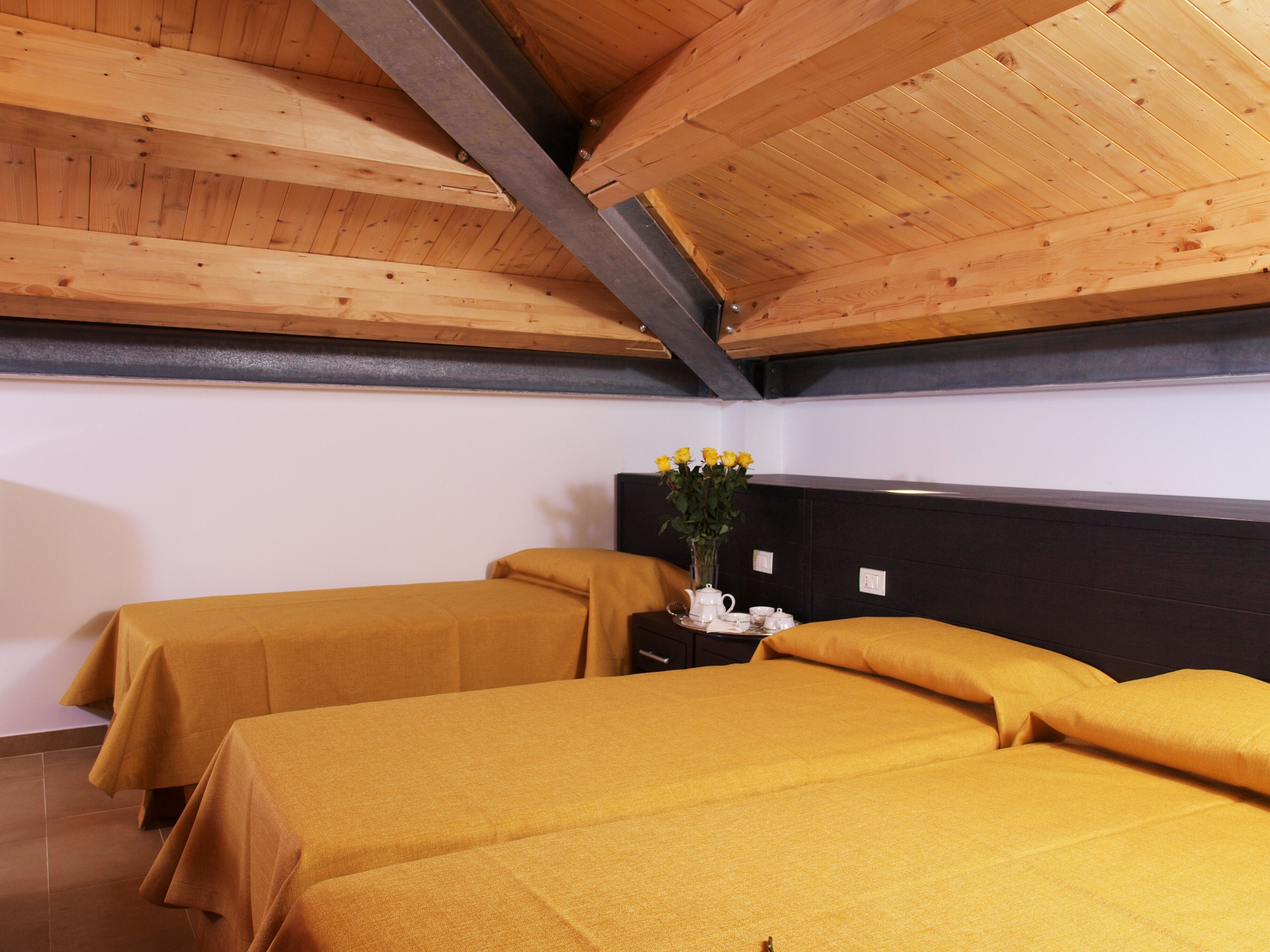 A room with two beds, wooden ceiling beams, a nightstand with a vase of flowers, and a black headboard.