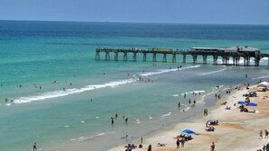A beach with a pier extending into the water, people swimming and sunbathing, and umbrellas on the sand. - Chairman of the Board at White Surf (Daytona Beach)