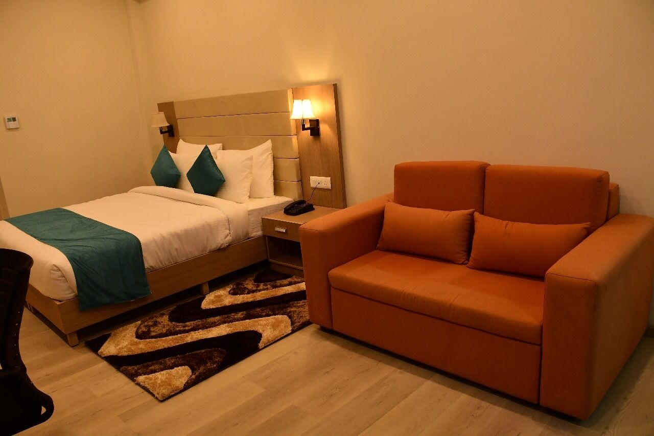 Una camera d'albergo con un letto, un divano arancione, un comodino e una lampada fissata al muro.