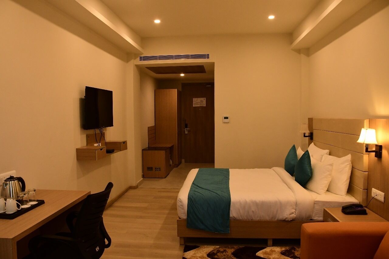 A hotel room with a bed, bedside table, lamp, TV, and a desk with a chair.