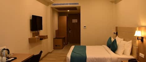 A hotel room with a bed, bedside table, lamp, TV, and a desk with a chair.