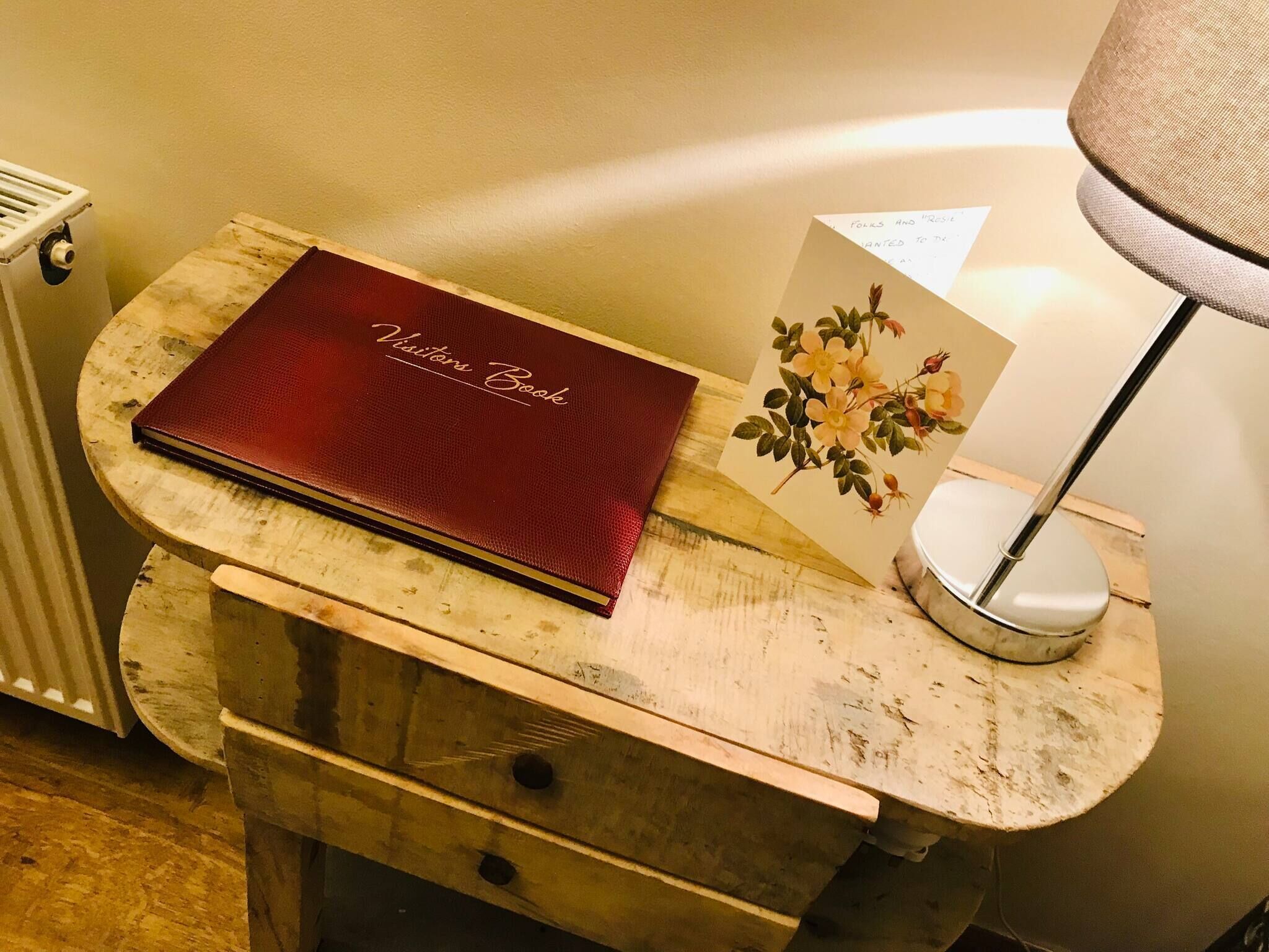 Een houten nachtkastje met een rood boek getiteld ‘Visitor Book’ en een kaartje met een bloemenmotief.