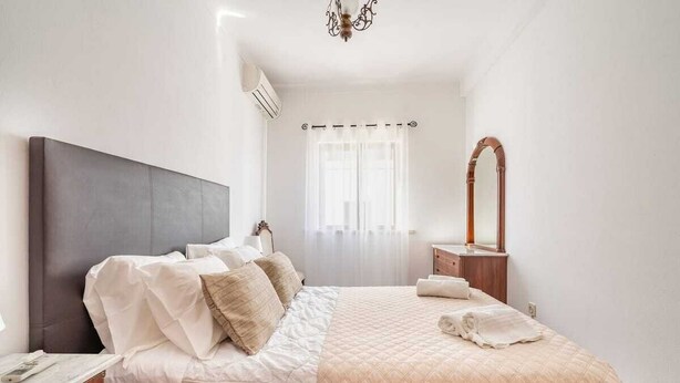 A neatly arranged bedroom with a large bed, a bedside table, a mirror, and a window with curtains. - Teixeira 's House (Tavira)
