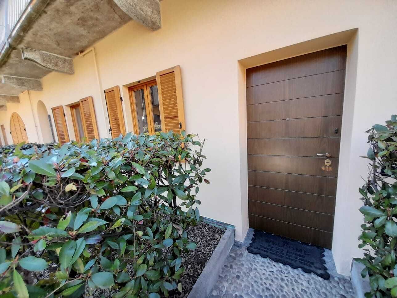 A building entrance with a wooden door, a small garden area with shrubs, and a concrete pathway leading to the door.