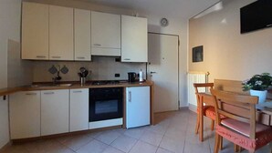 A compact kitchen with white cabinets, a stove, and a small refrigerator. There is a wooden dining table with chairs and a potted plant on the right.