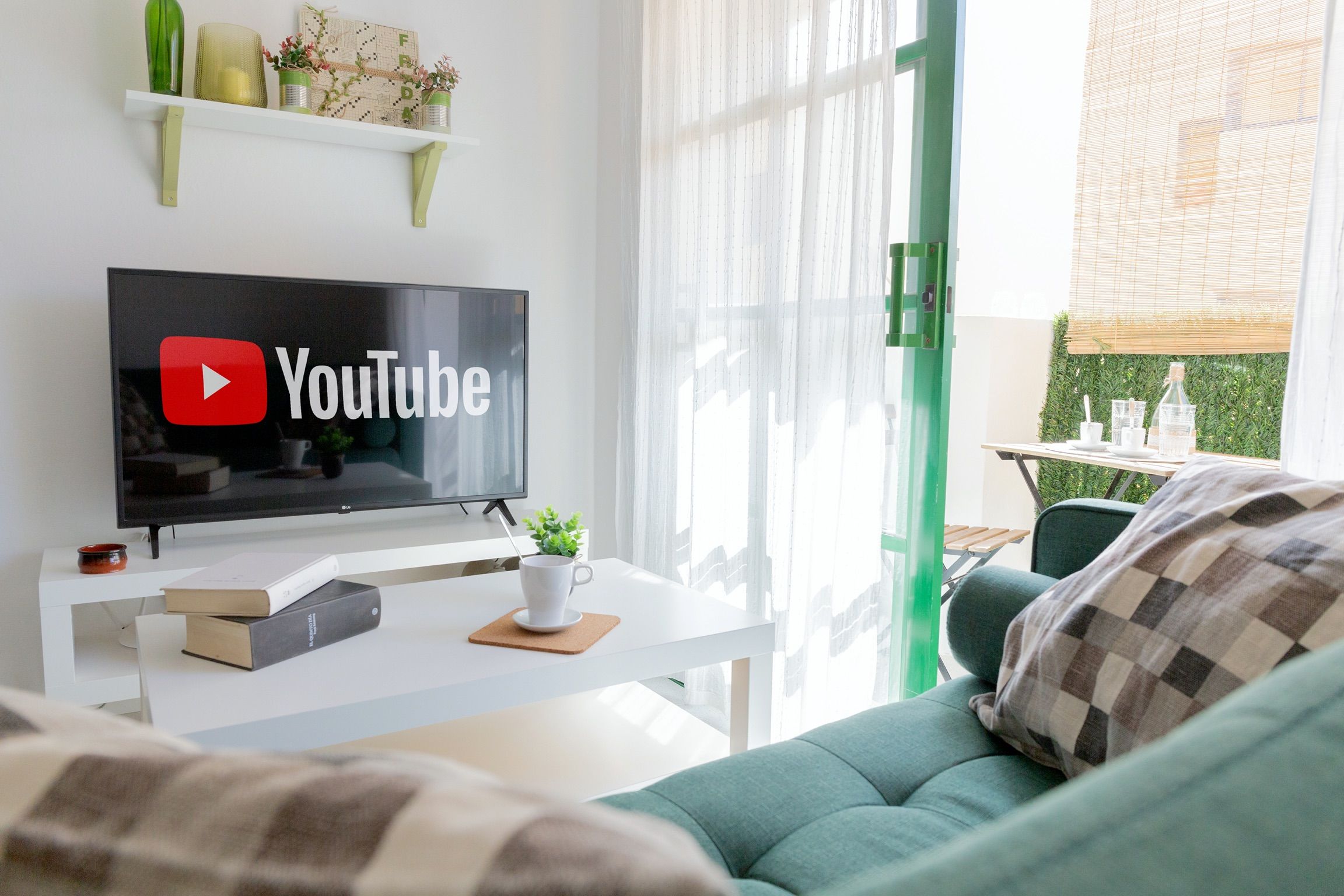 A modern living room with a flat-screen TV displaying YouTube, a white coffee table with a cup and a book, a green sofa with a plaid throw, and a window with sheer curtains.