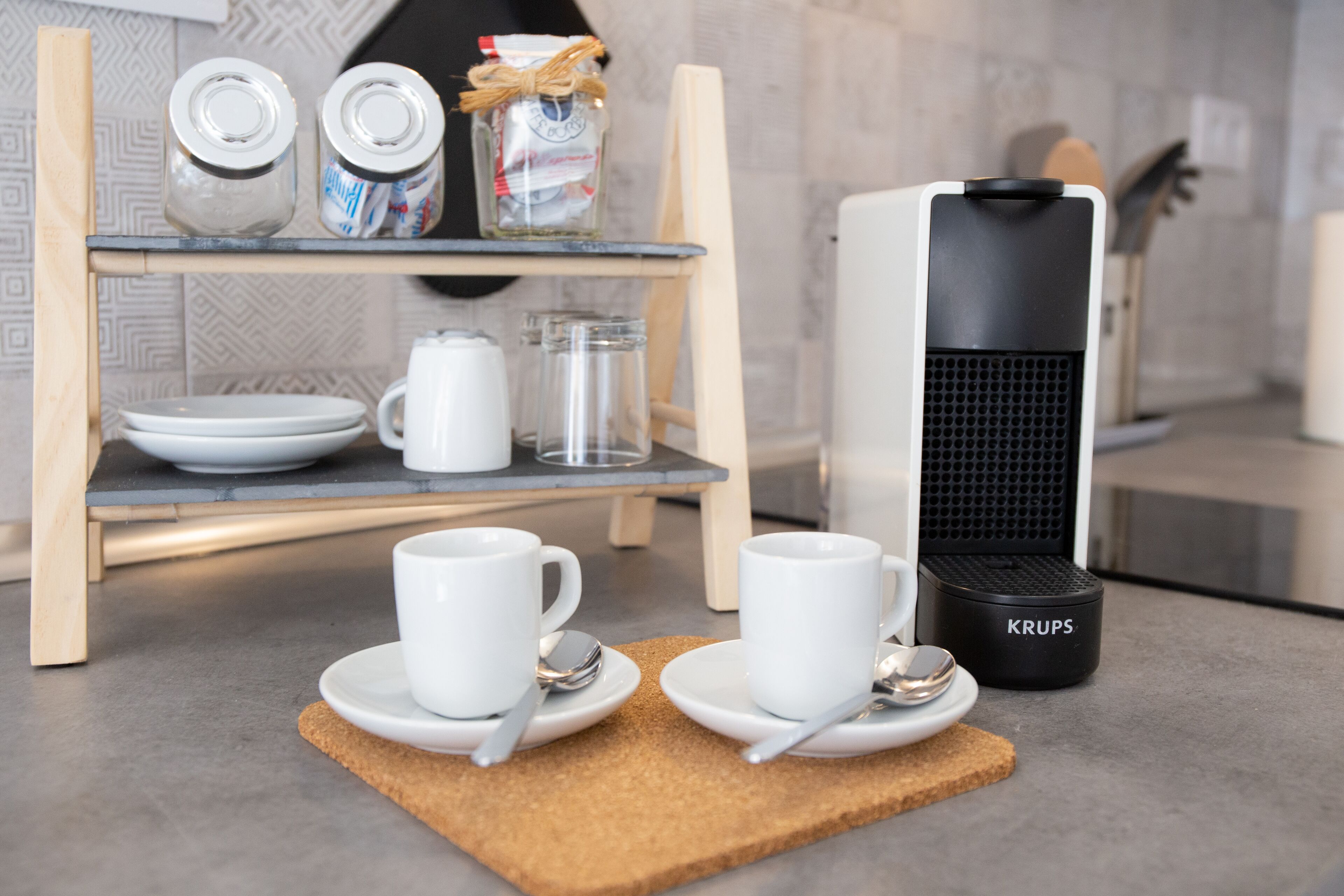 A modern kitchen with a coffee maker, two cups, a jar, and a shelf with plates and glasses.