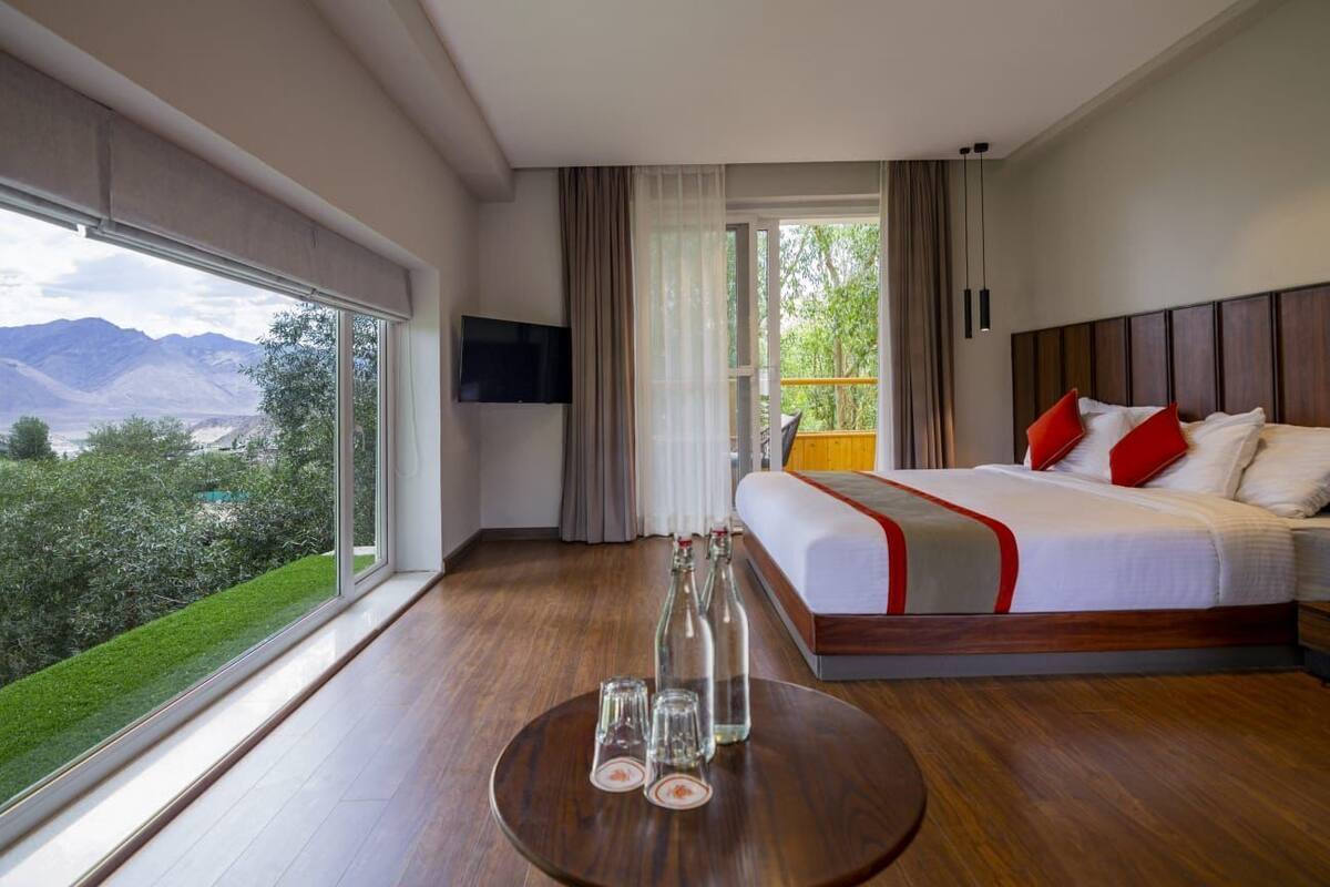 A hotel room with a large bed, a TV, a balcony with a view of mountains, and a round table with glasses.