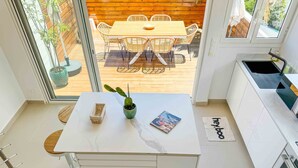 A modern kitchen with a white countertop, a small potted plant, and a magazine. There is a dining area with a wooden table and chairs visible through the glass door.
