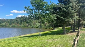 Un parque tranquilo junto al lago, con valla de madera, abundante vegetación y un cielo azul despejado.