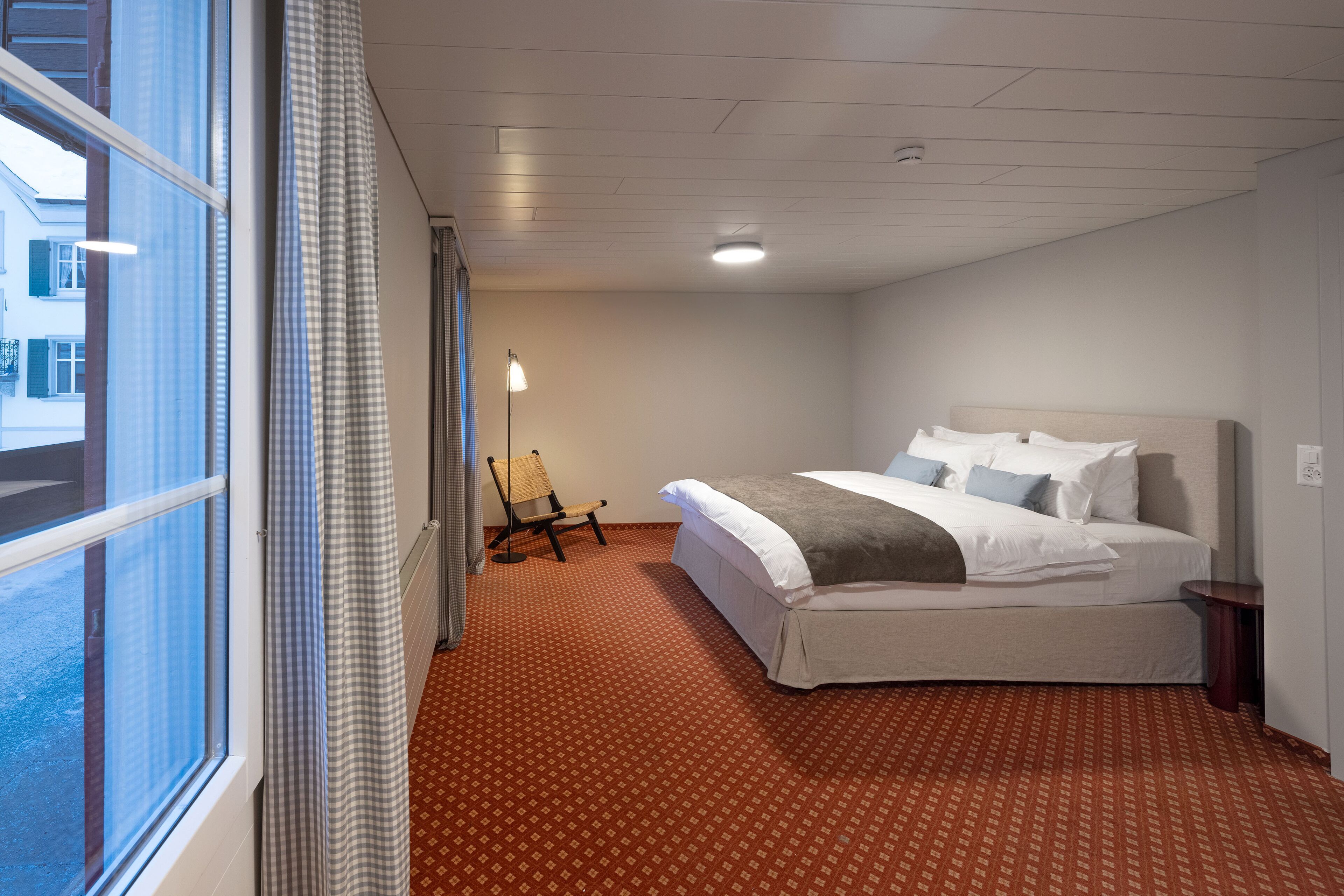 A hotel room with a large window, a bed with white linens, a chair, and a floor lamp.