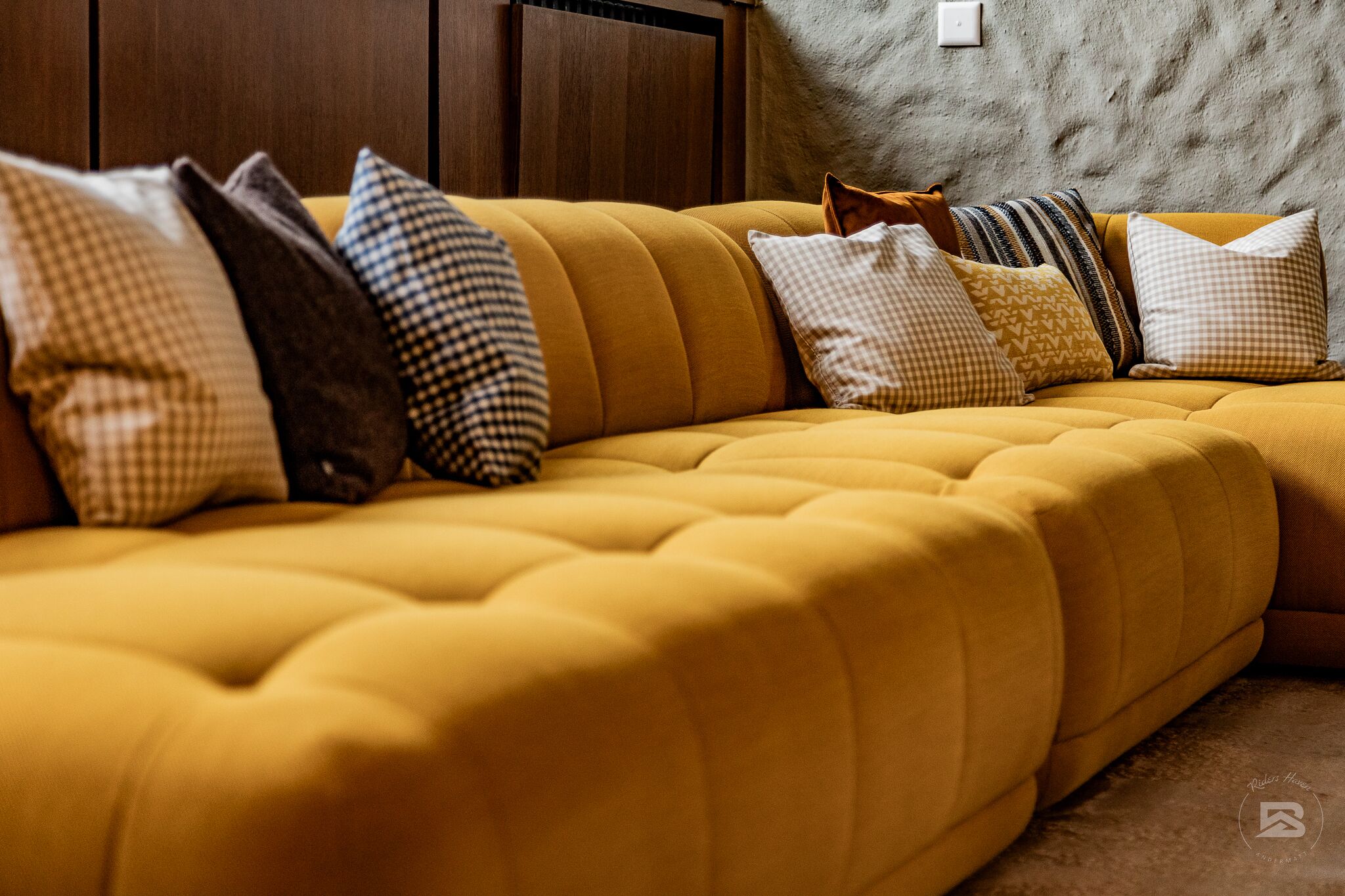 Ein gelbes Sofa mit vielen Kissen in einem Raum mit Betonwand und Holzvertäfelung.