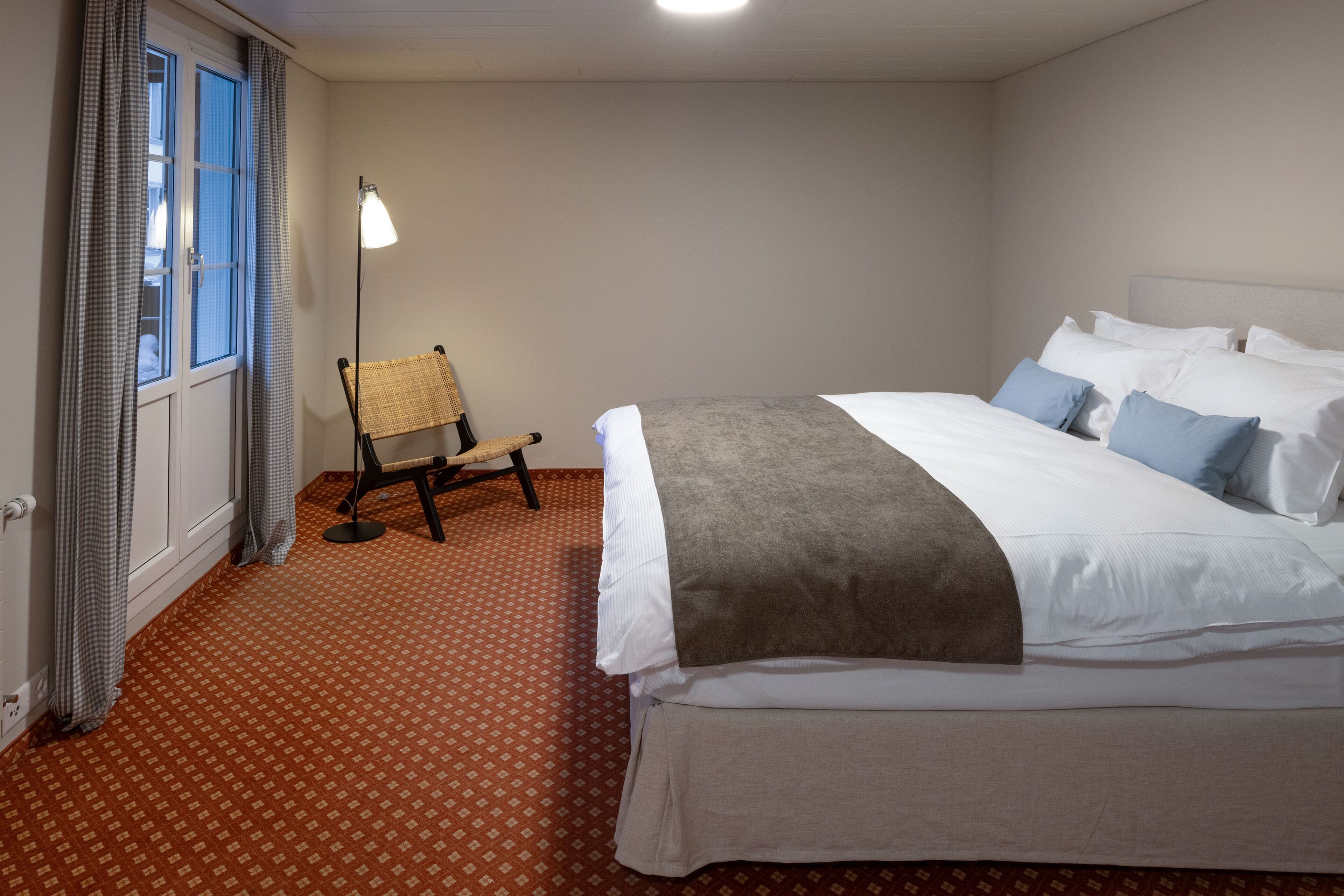 A hotel room with a bed, a chair, a lamp, and a window with curtains.