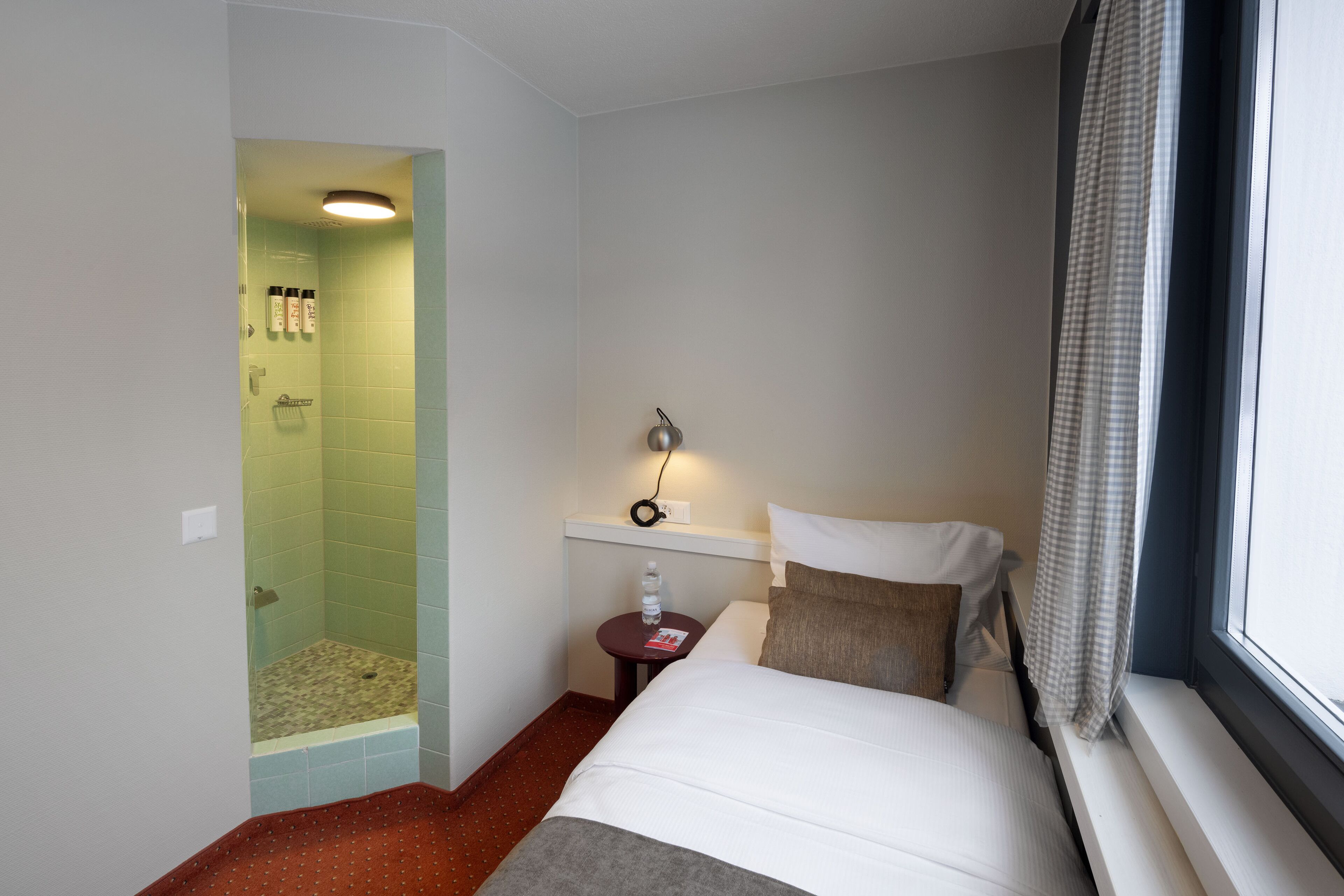 Une chambre d’hôtel avec un lit, une petite table sur laquelle se trouvent une bouteille et un livre, une table de chevet avec une lampe, une fenêtre avec un rideau et un espace douche avec un porte-serviettes.
