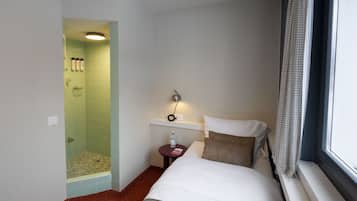 Une chambre d’hôtel avec un lit, une petite table sur laquelle se trouvent une bouteille et un livre, une table de chevet avec une lampe, une fenêtre avec un rideau et un espace douche avec un porte-serviettes.
