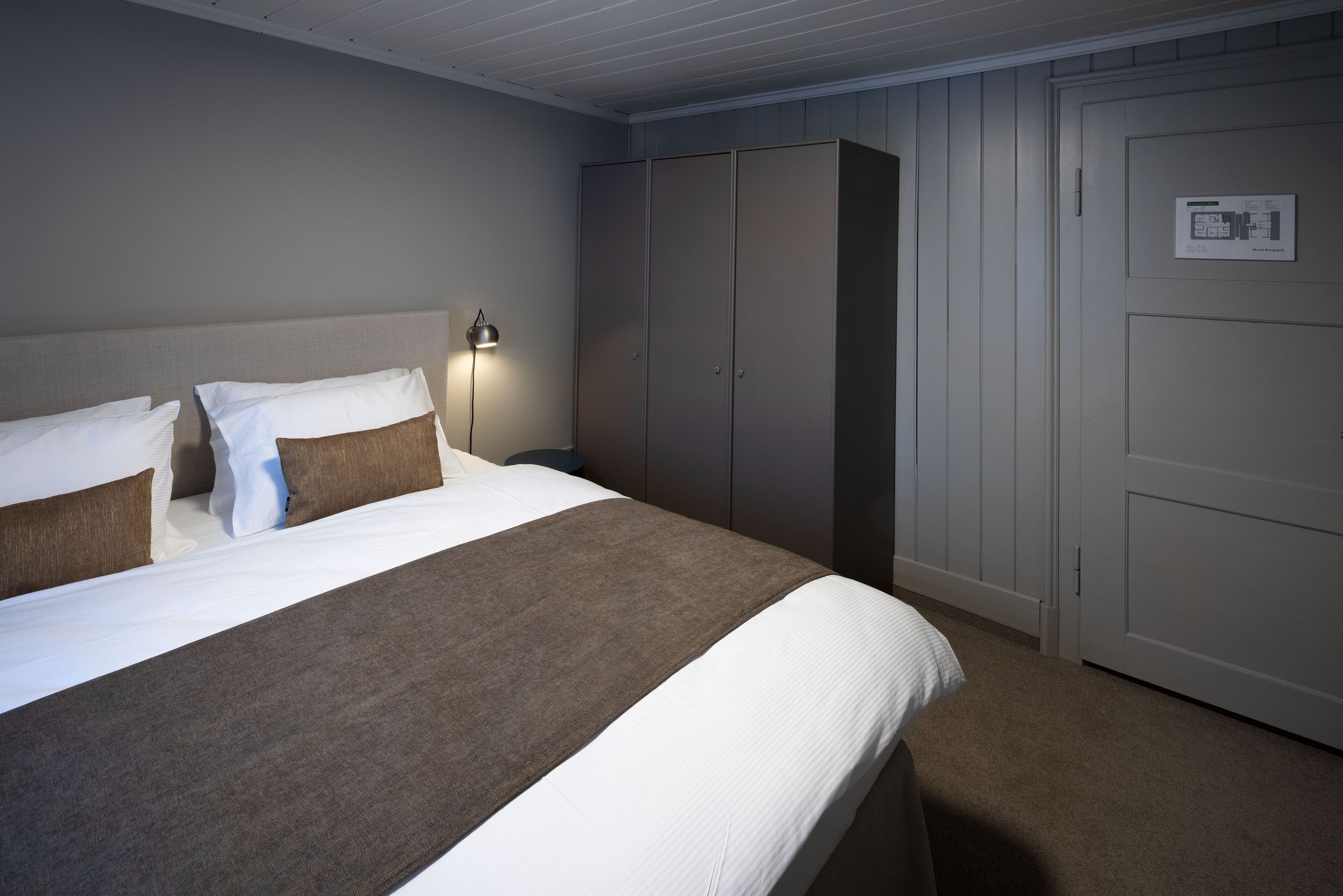 Ein ordentlich bezogenes Bett mit weißen und braunen Textilien, eine Nachttischlampe und ein Kleiderschrank mit Bedienfeld.