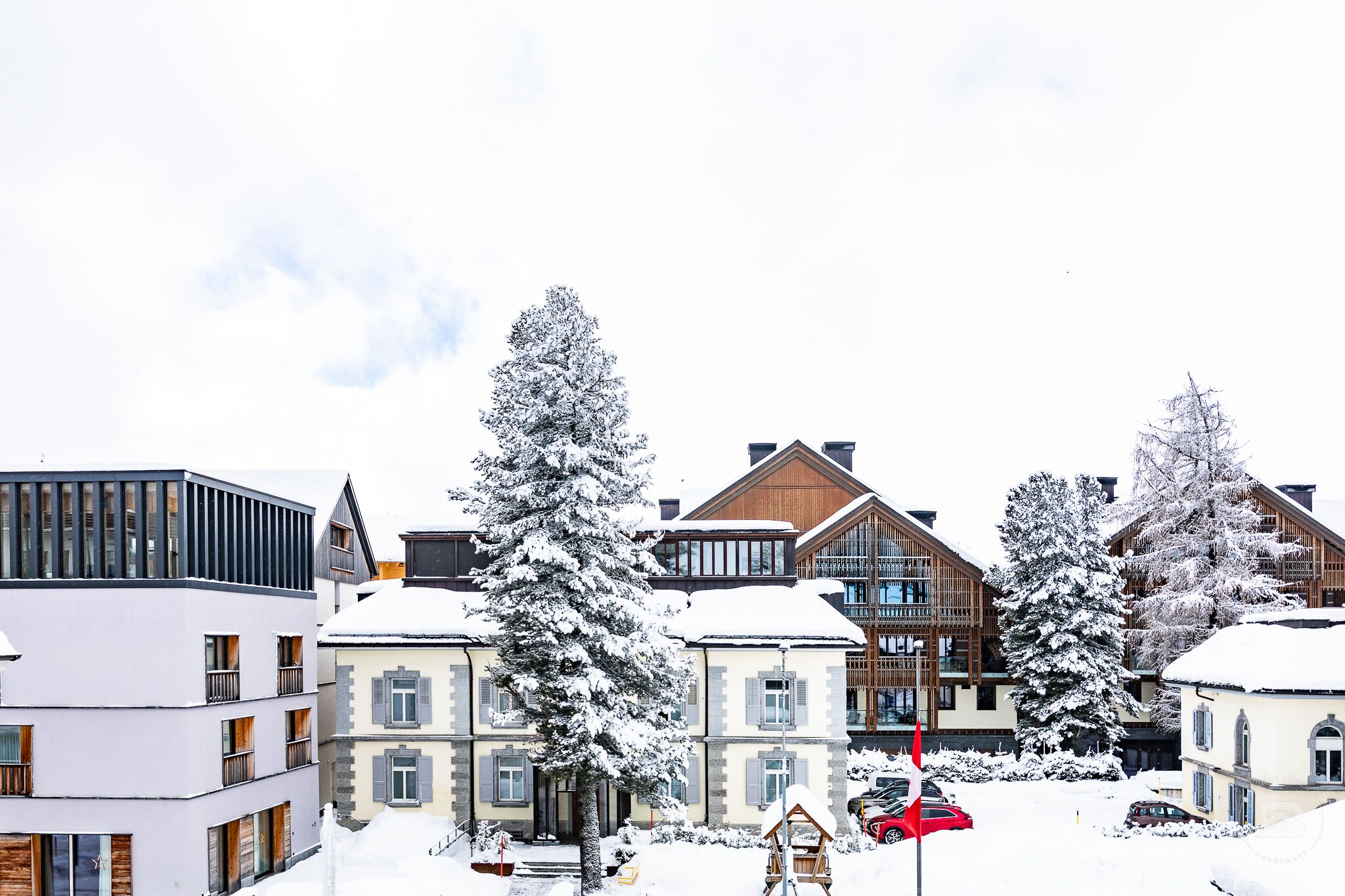 Een besneeuwde woonwijk met moderne gebouwen, waaronder een meerlaags gebouw met balkons en een chalet in traditionele stijl met een houten gevel.