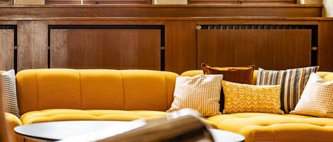 A yellow sofa with patterned cushions in a room with wooden paneling and a window with a geometric pattern.