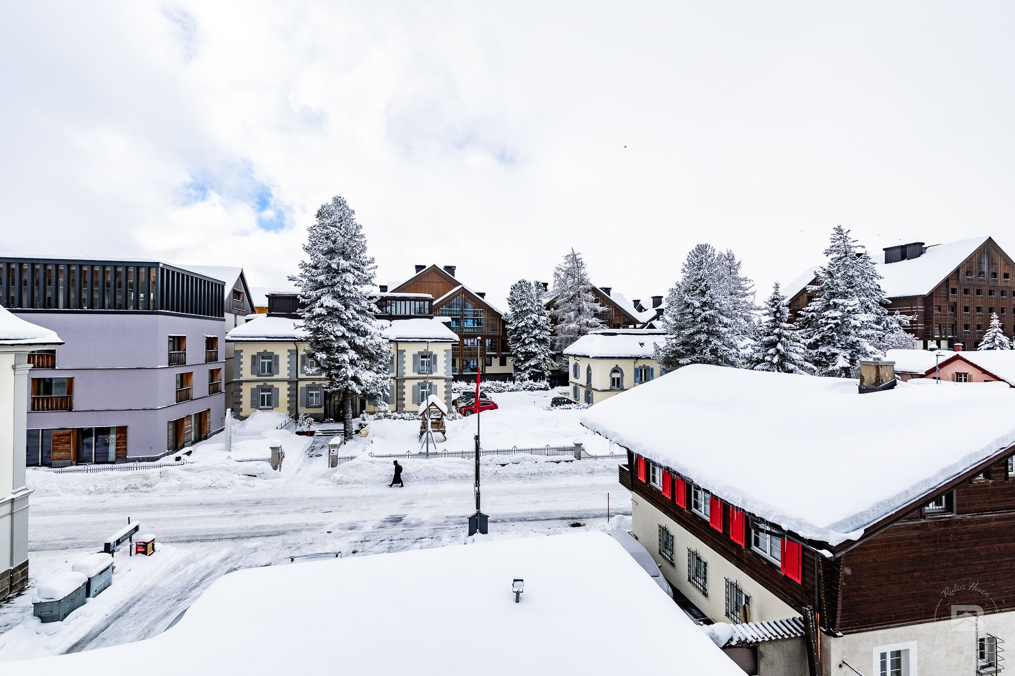Une ville enneigée, avec des bâtiments, dont un chalet en bois aux volets rouges.