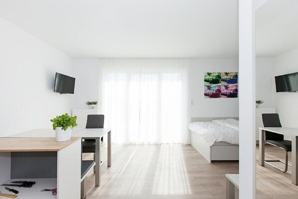 A modern, minimalist hotel room with a wooden desk, a bed, a TV, and a large window with sheer curtains.