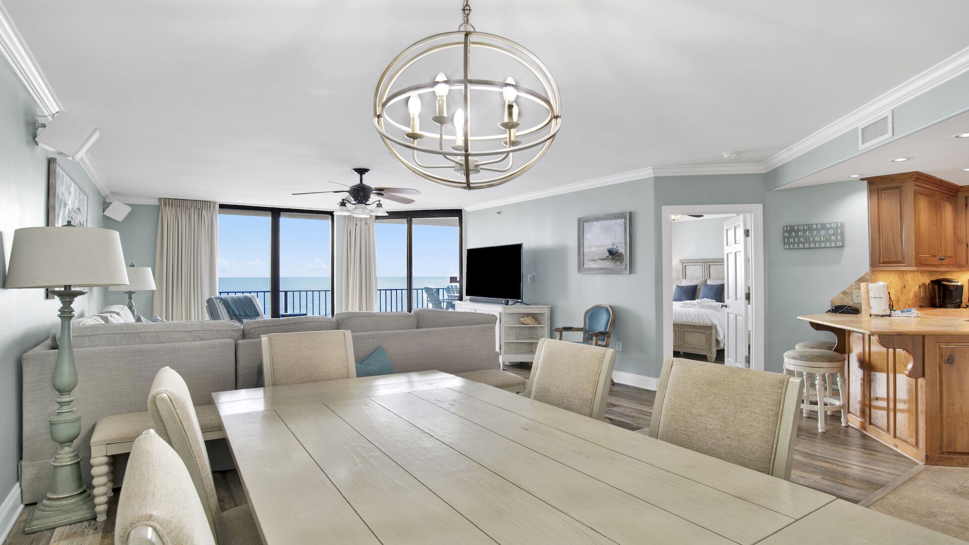 Un salon spacieux avec une grande table à manger, un ventilateur de plafond et une vue sur l’océan grâce à de grandes fenêtres.
