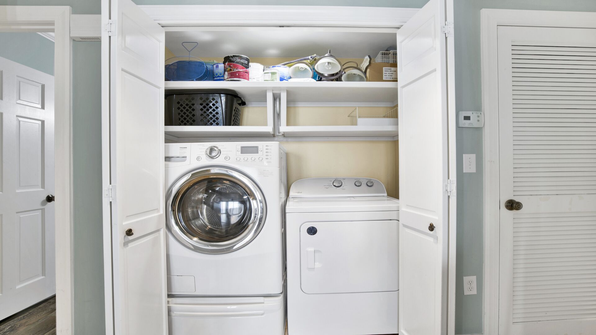 A compact laundry room with a front-loading washer and dryer, a shelf above the washer, and a door with a louvered window.