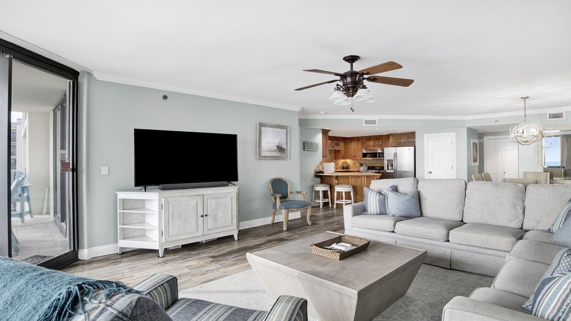 A spacious living room with a flat-screen TV, a ceiling fan, and a kitchen area in the background.
