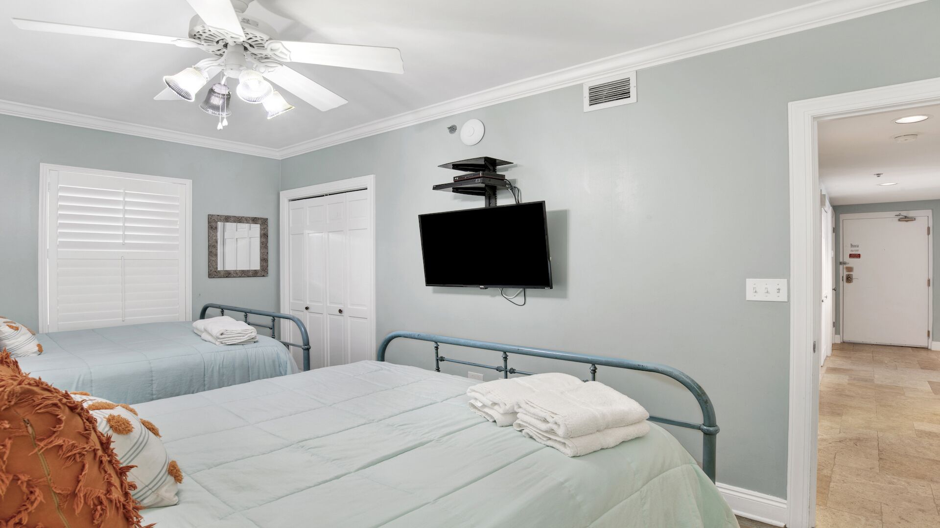A hotel room with two beds, a ceiling fan, a TV mounted on the wall, and a door leading to another room.