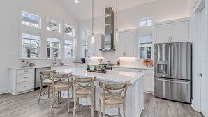 Una cocina moderna con muebles blancos, una isla grande con taburetes, electrodomésticos de acero inoxidable e iluminación colgante.