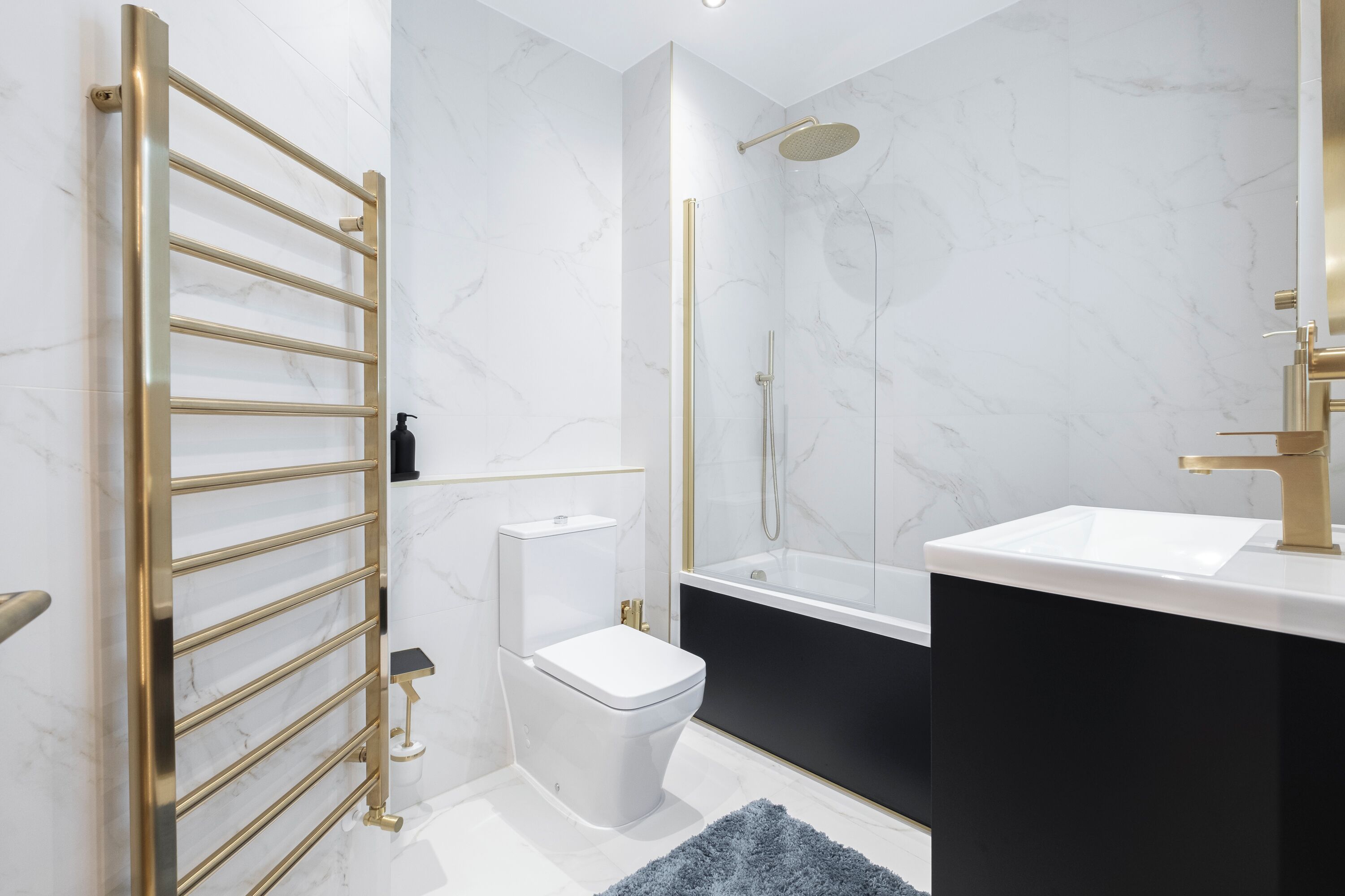 A modern bathroom with a gold towel rack, a white toilet, a black bathtub, and a white sink with a gold faucet.
