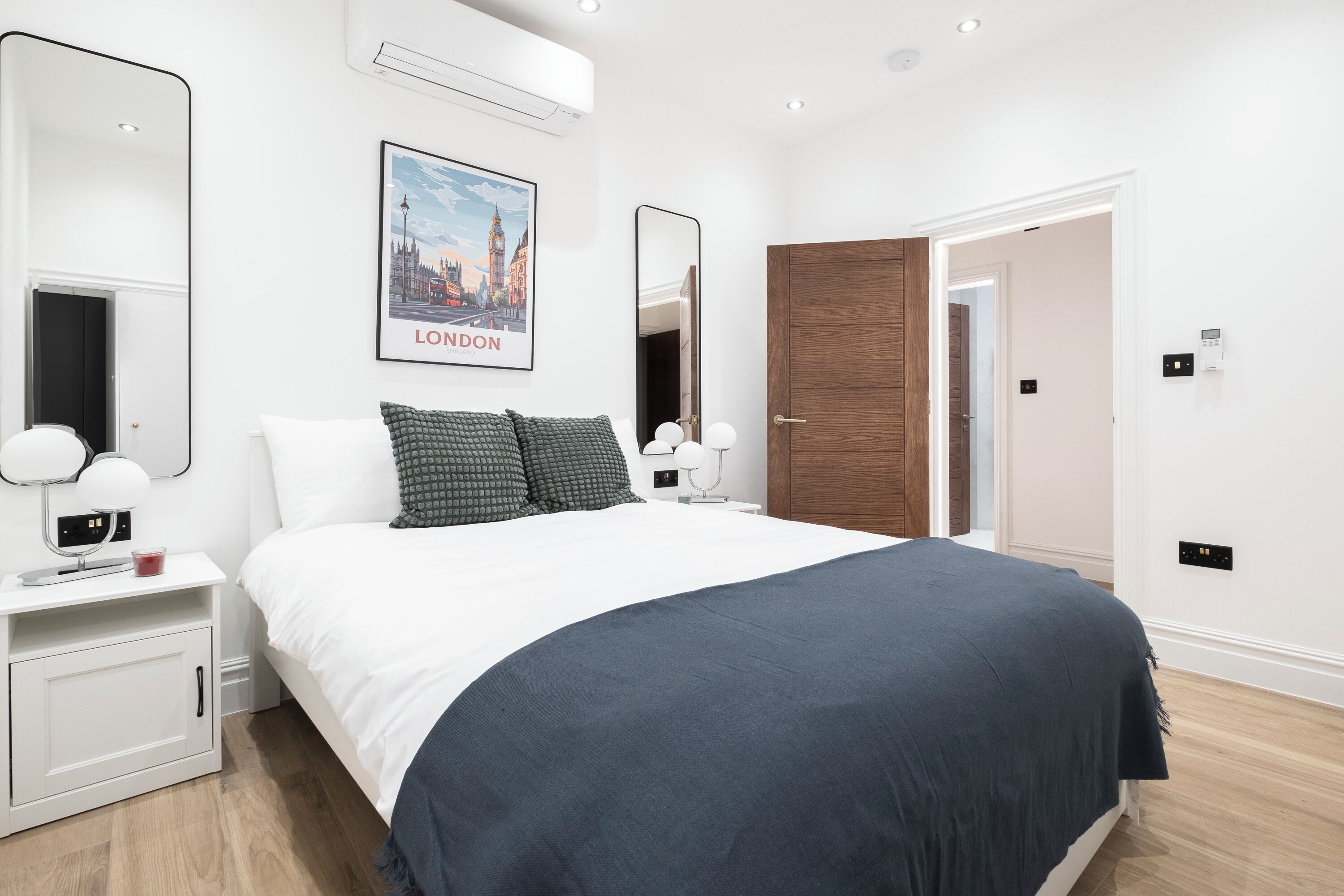 Ein modernes Schlafzimmer mit einem großen Bett, weißen Nachttischen, einem Spiegel und einem gerahmten Bild von London.