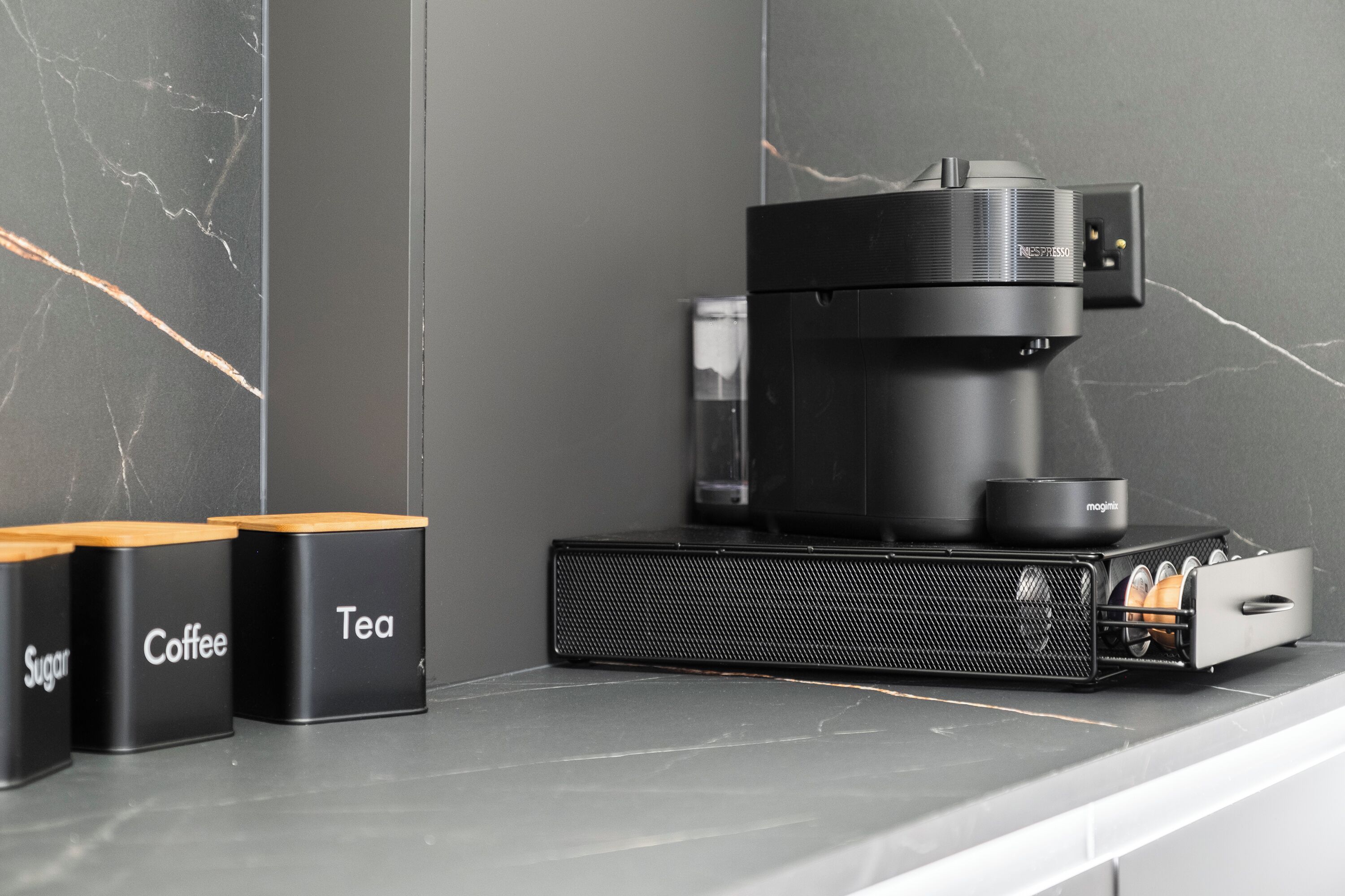 Una cocina con cafetera, recipientes etiquetados para azúcar, café y té, y un salpicadero de mármol.