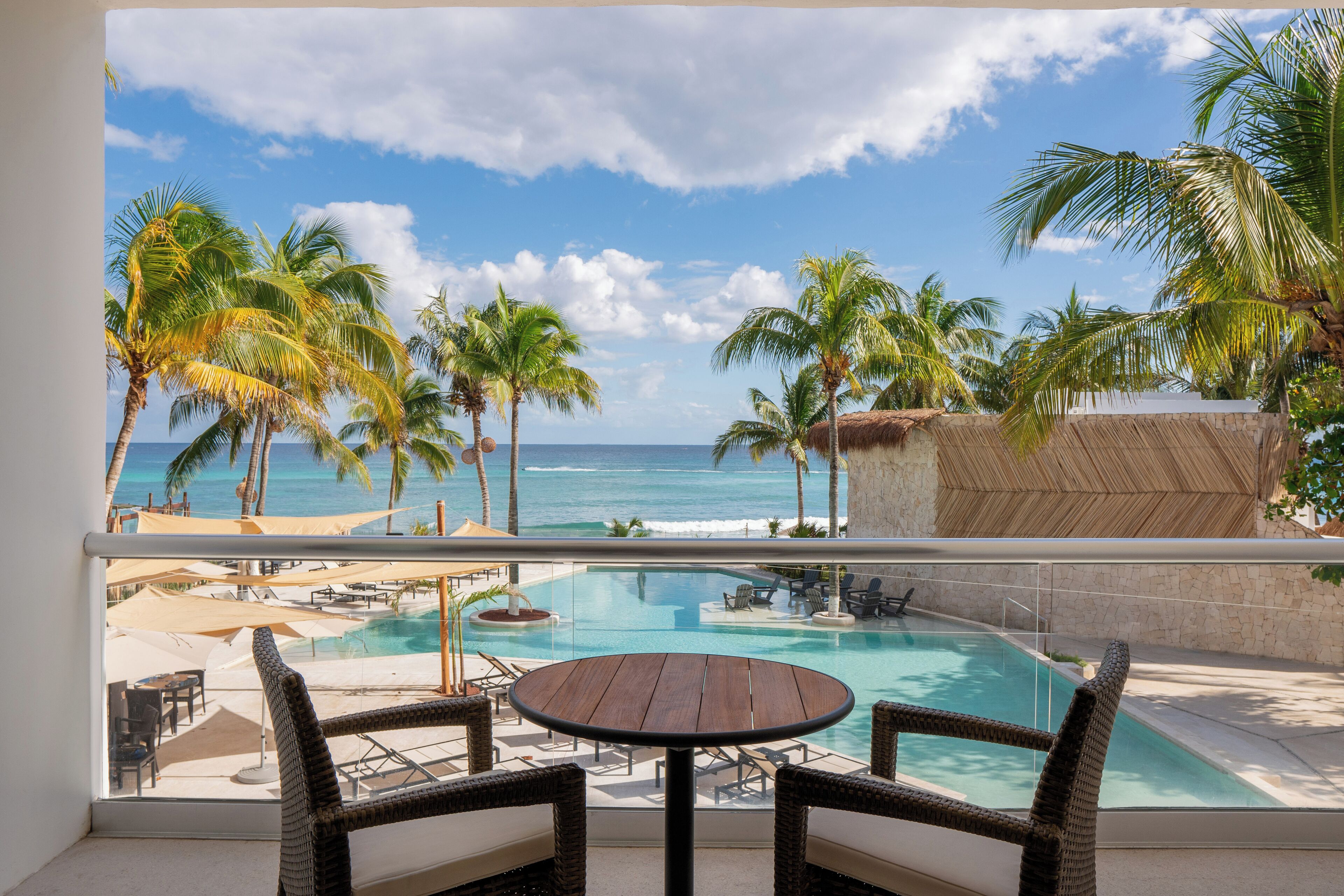 Un espace aménagé au bord de la piscine, avec des fauteuils en osier, une table en bois et une vue sur l’océan et les palmiers.