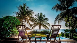 Two wooden lounge chairs facing a pool with a view of the ocean and palm trees.
