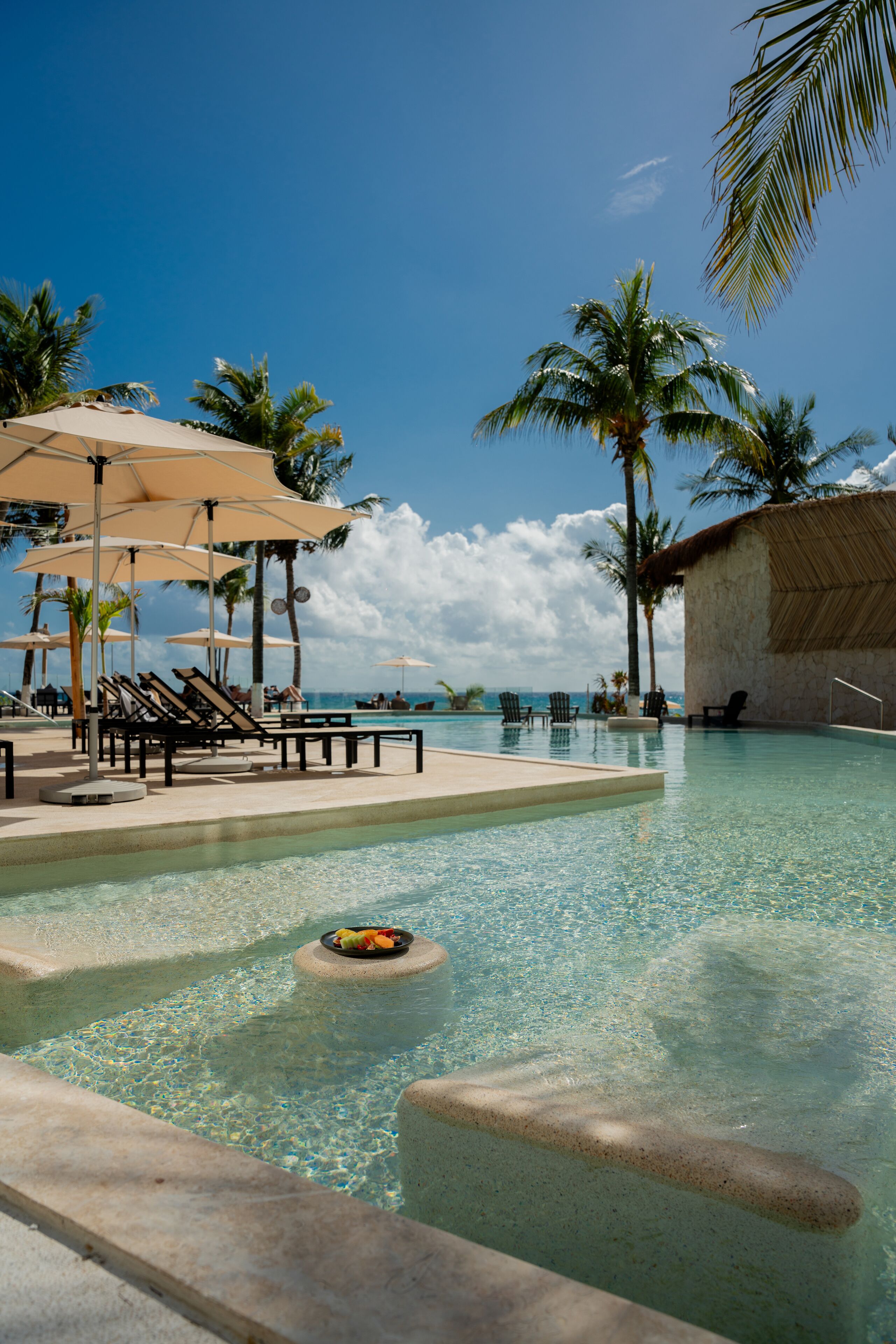 A clear pool with lounge chairs and umbrellas, surrounded by palm trees.